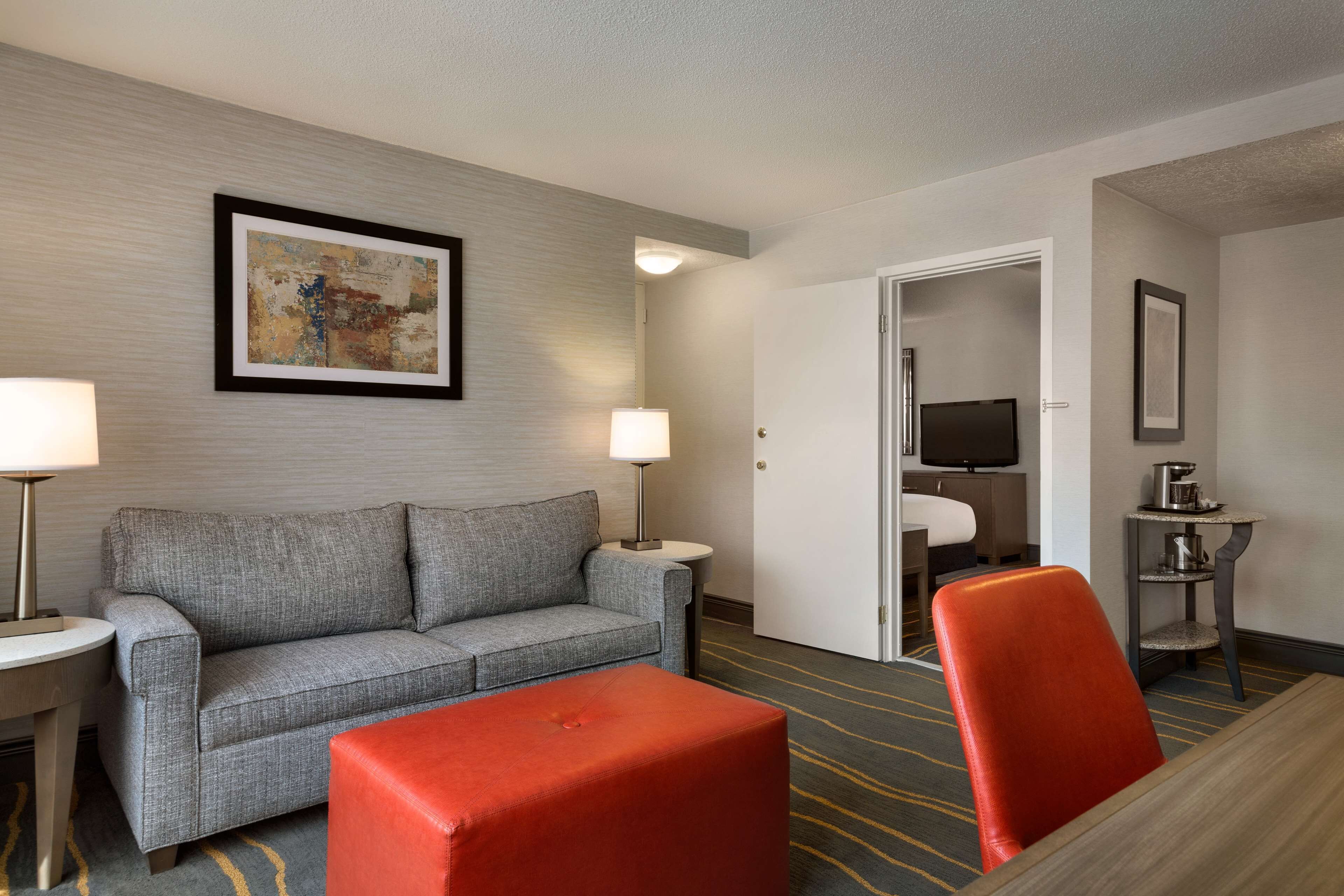 Una camera d'albergo con un divano grigio, un poggiapiedi rosso, un tavolino con una lampada, una TV e un quadro appeso al muro.