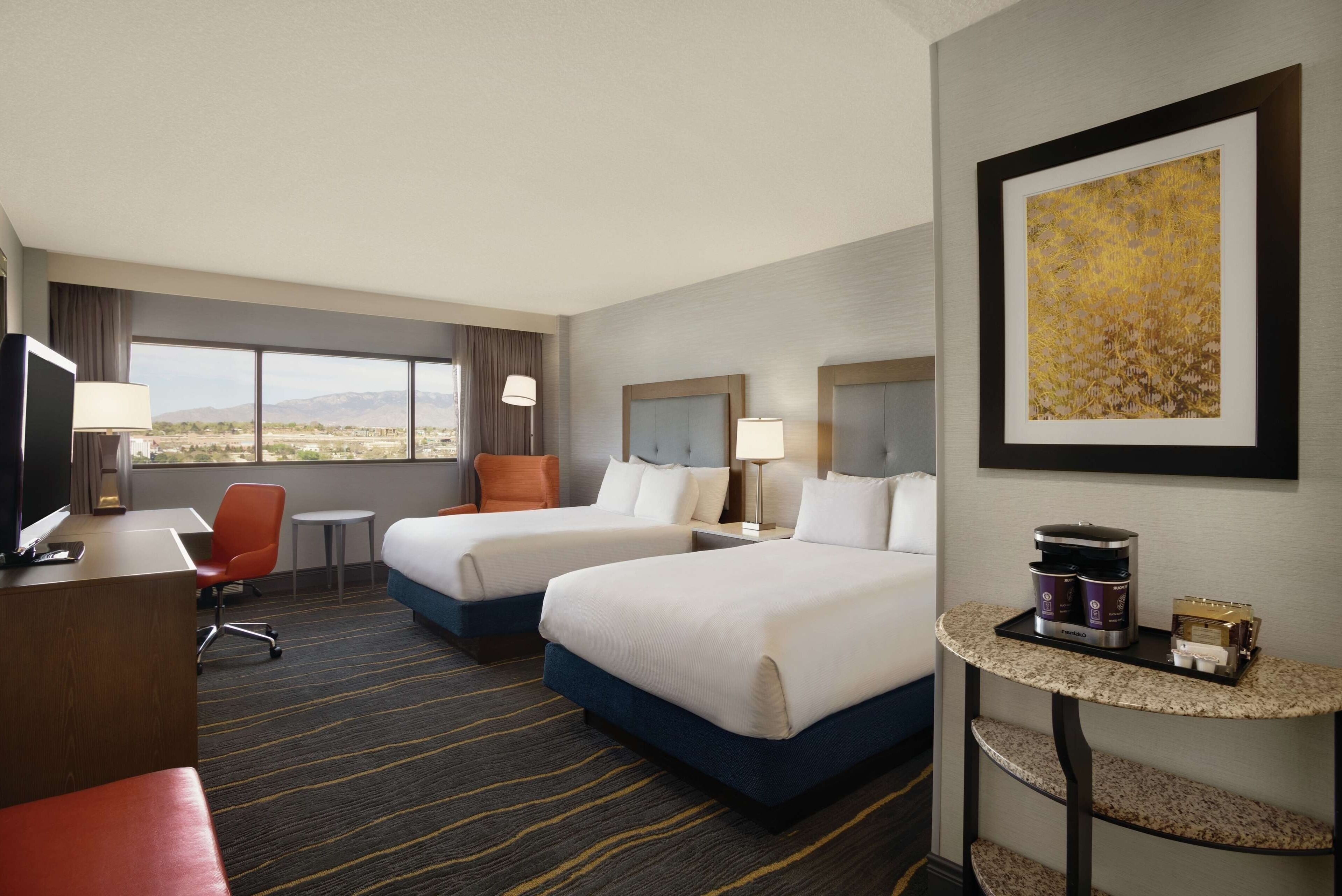 A hotel room with two beds, a desk with a computer, a chair, a small table with a coffee maker, and a large window with a view of the mountains.