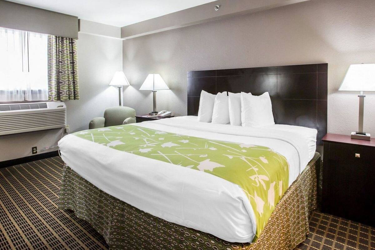 A hotel room with a bed, two bedside lamps, a nightstand, a telephone, a window with curtains, and an air conditioning unit.
