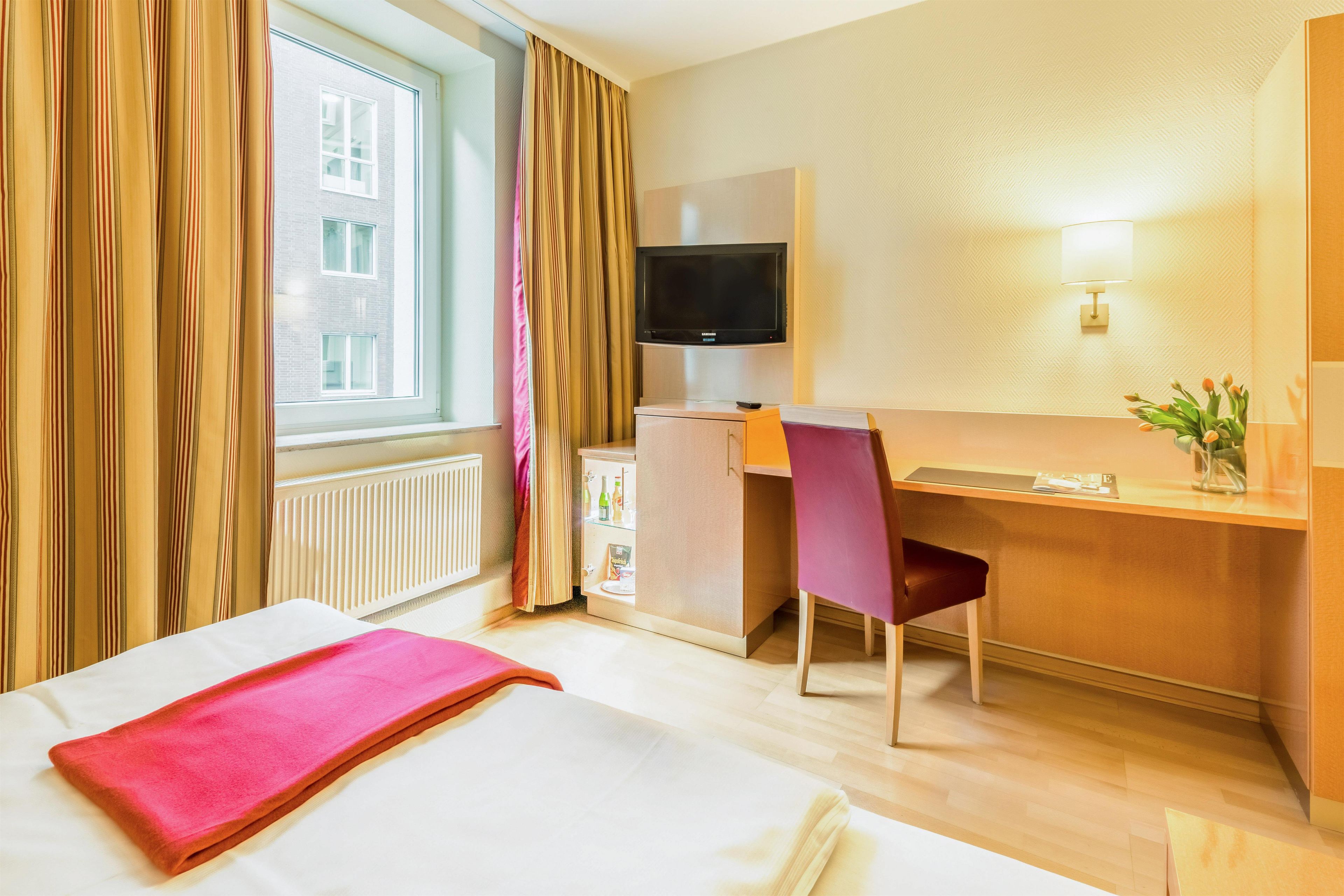 A hotel room with a bed, a desk with a chair, a television, and a window with curtains.