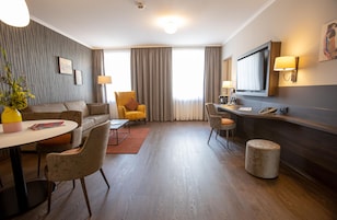 Una habitación de hotel moderna con sofá, sillones, una mesa de comedor, televisión y escritorio con lámpara.
