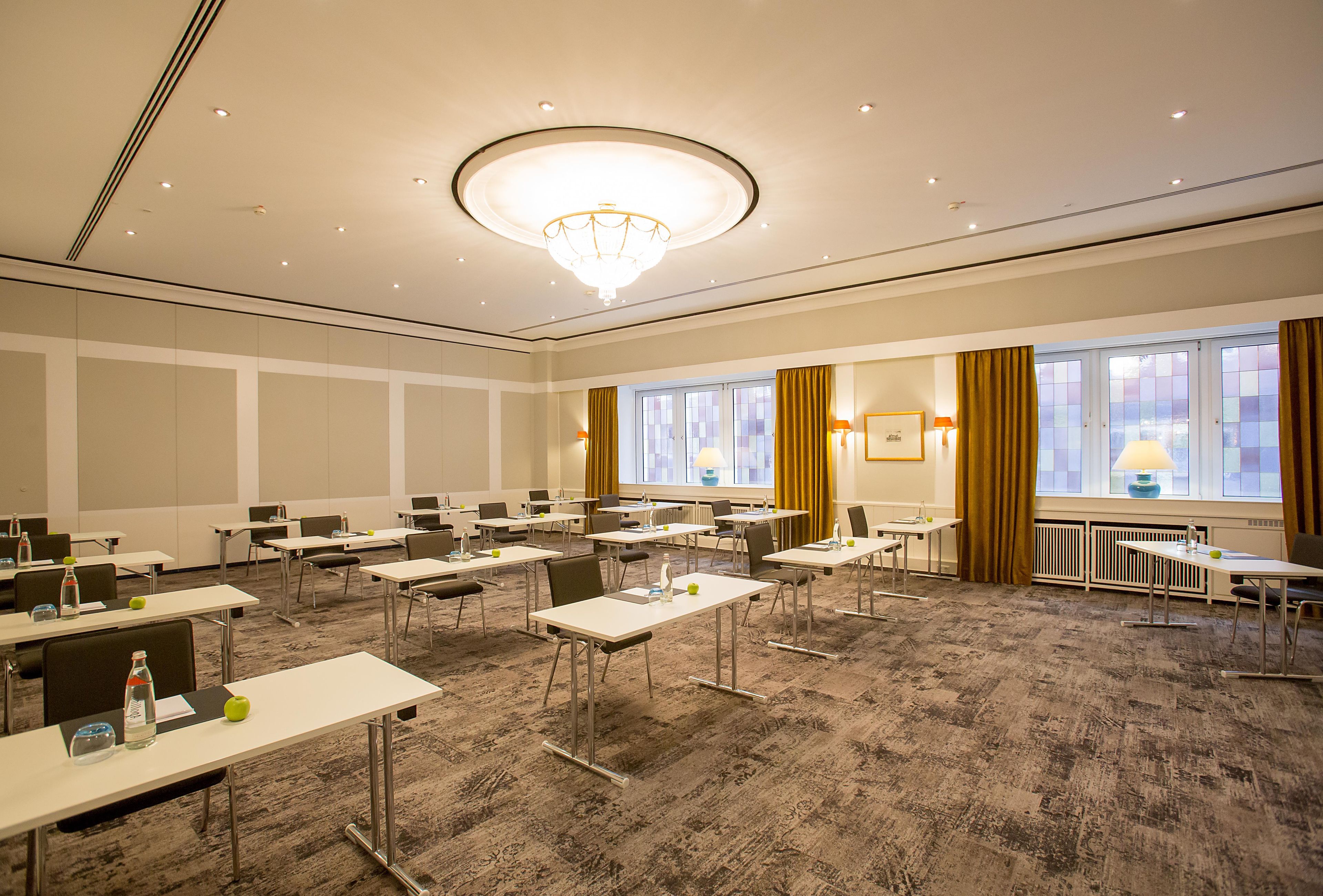 Un salón de conferencias con mesas, sillas, una lámpara de araña y ventanas con cortinas.
