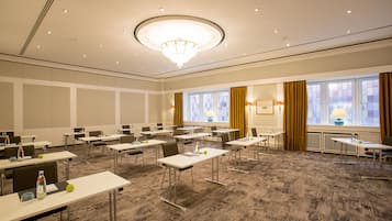 Un salón de conferencias con mesas, sillas, una lámpara de araña y ventanas con cortinas.