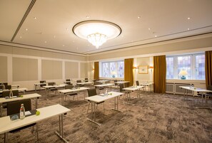 Un salón de conferencias con mesas, sillas, una lámpara de araña y ventanas con cortinas.