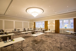 Un salón de conferencias con mesas, sillas, una lámpara de araña y ventanas con cortinas.