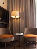 Habitación de hotel con dos sillones, una lámpara de pie, una mesita con un libro y un televisor montado en la pared.