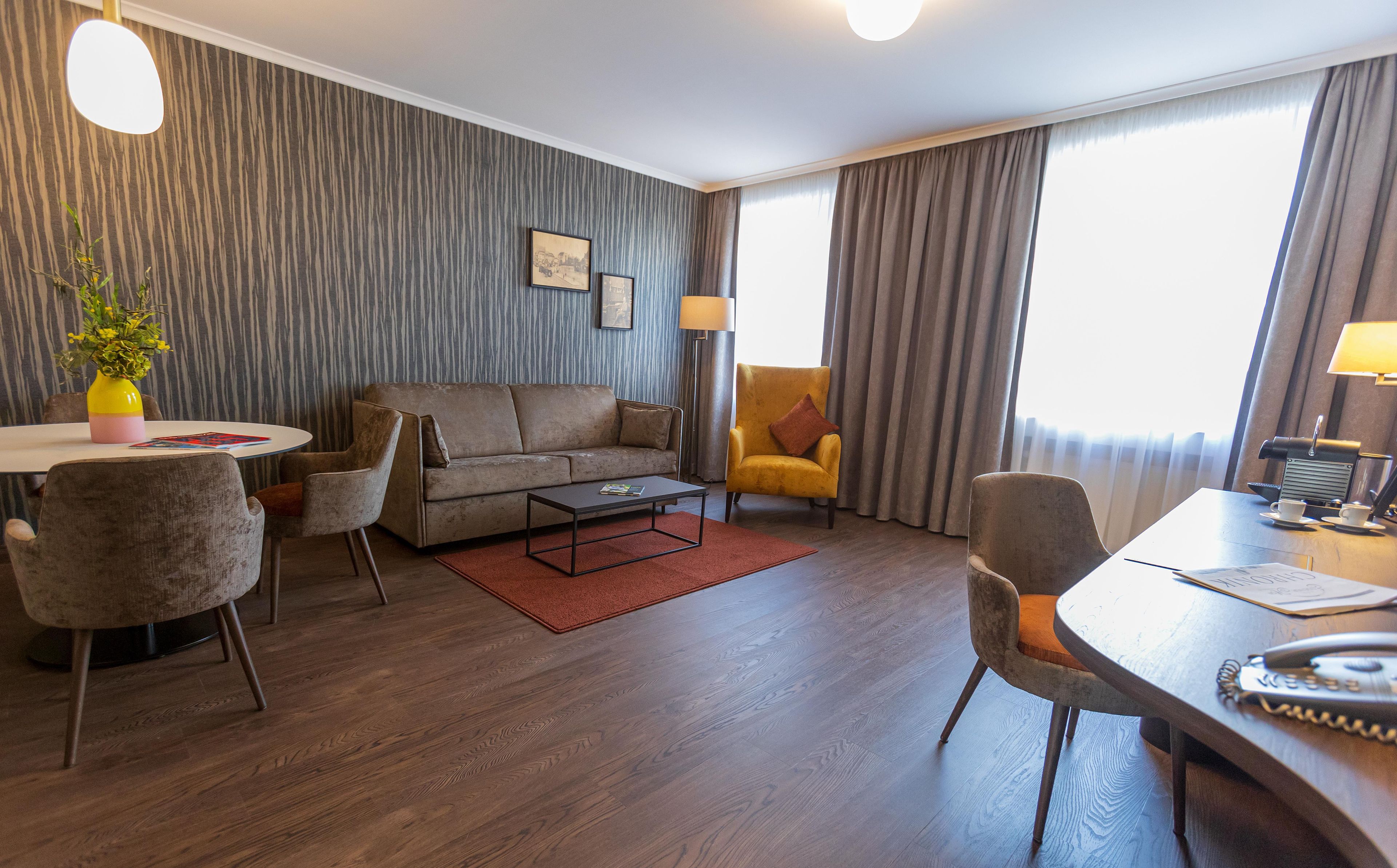 A hotel room with a sofa, armchairs, a coffee table, a dining table, and a lamp.