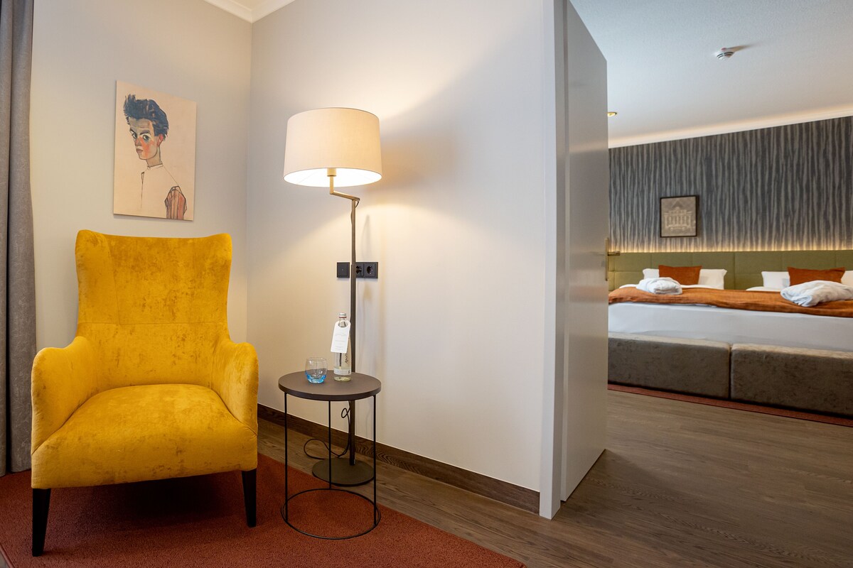 Een hotelkamer met een gele fauteuil, een nachtkastje met een lamp en een bed met hoofdeinde.