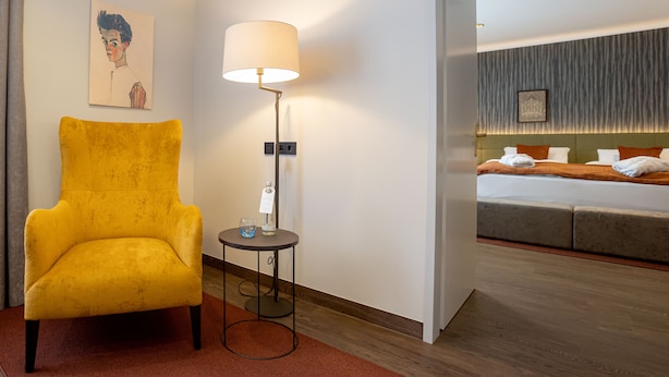 A hotel room with a yellow armchair, a bedside table with a lamp, and a bed with a headboard.