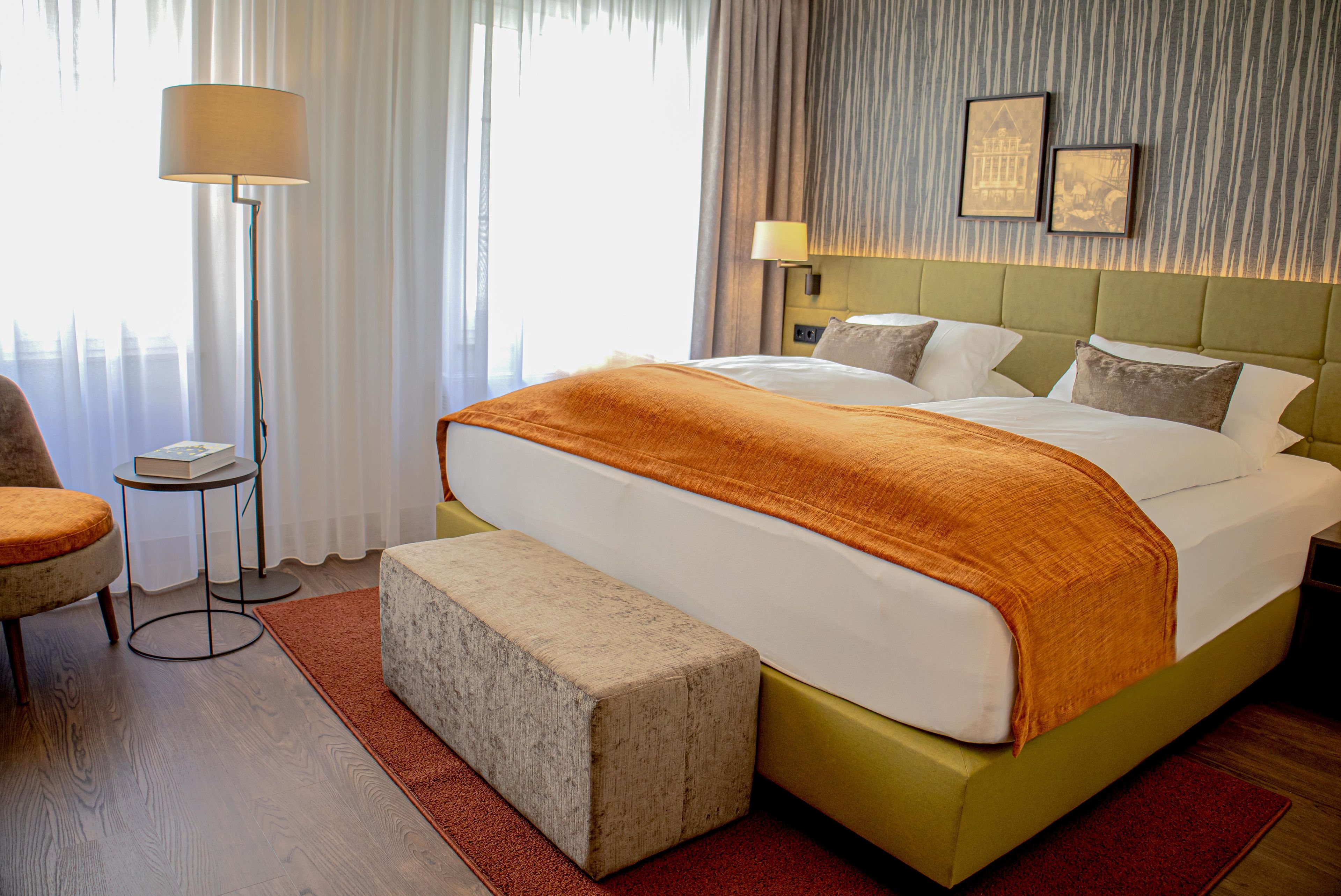 A hotel room with a large bed, a bedside table, a chair, a floor lamp, and a view of the city.