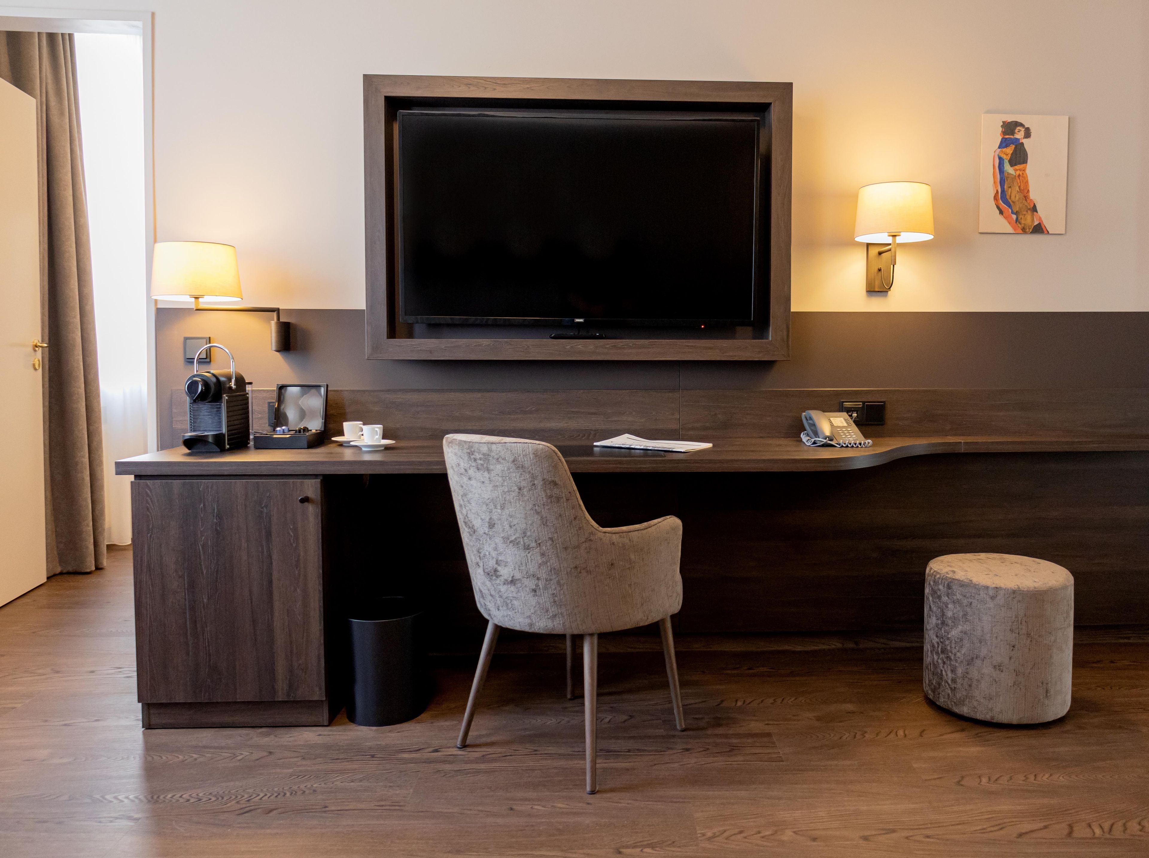 Ein Hotelzimmer mit einem großen Flachbildfernseher an der Wand, einem Schreibtisch mit Stuhl, einer Lampe und einer kleinen Grafik an der Wand.