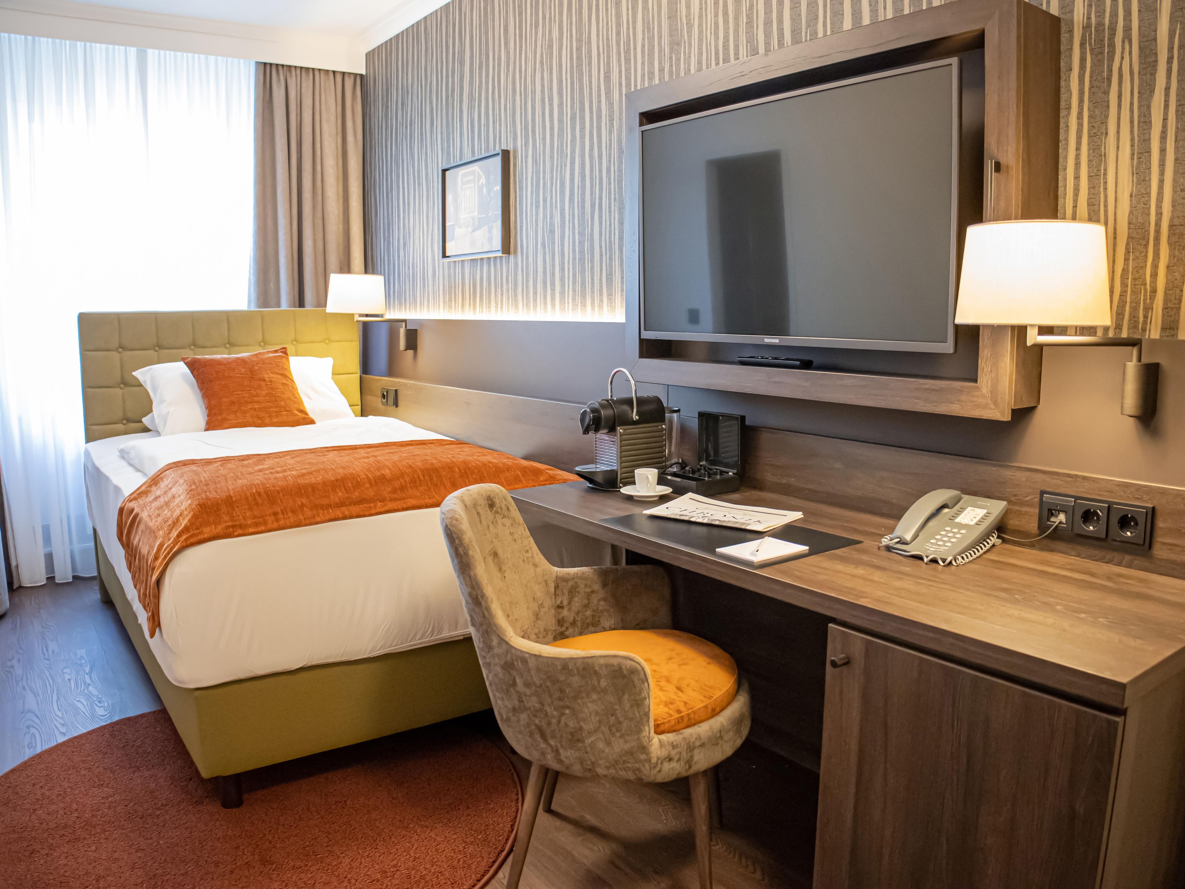 Habitación de hotel con cama, escritorio, silla, televisor, lámpara y teléfono.