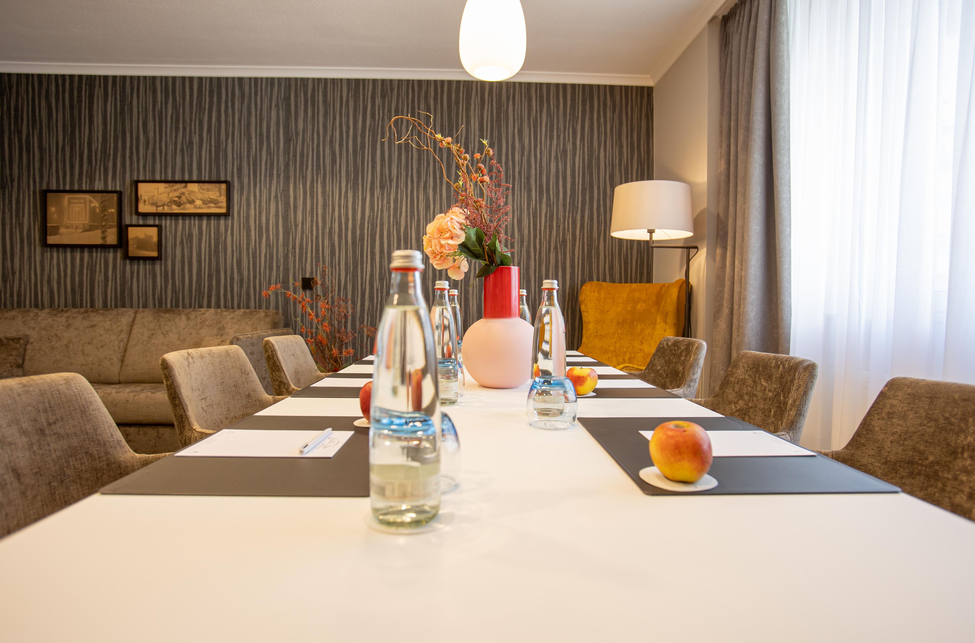 A hotel room with a dining table set for six, a sofa, a lamp, and framed pictures on the wall.
