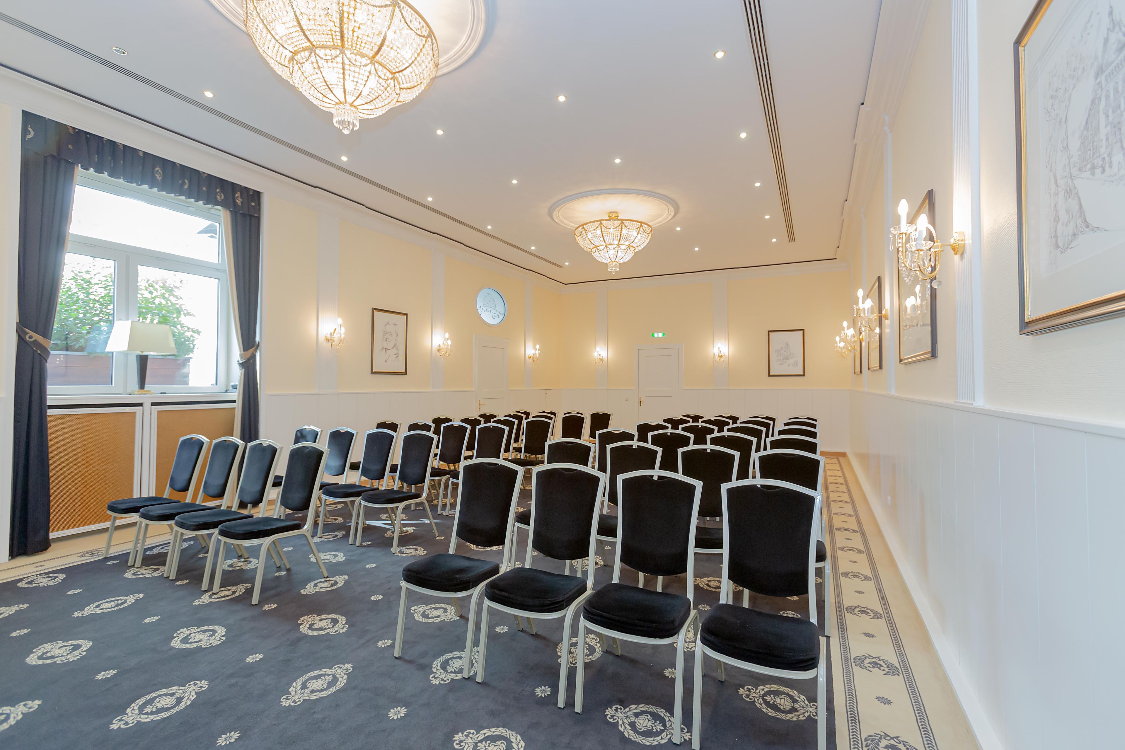 Un salón de conferencias con filas de sillas, un candelabro, obras de arte enmarcadas y un ventanal con cortinas.