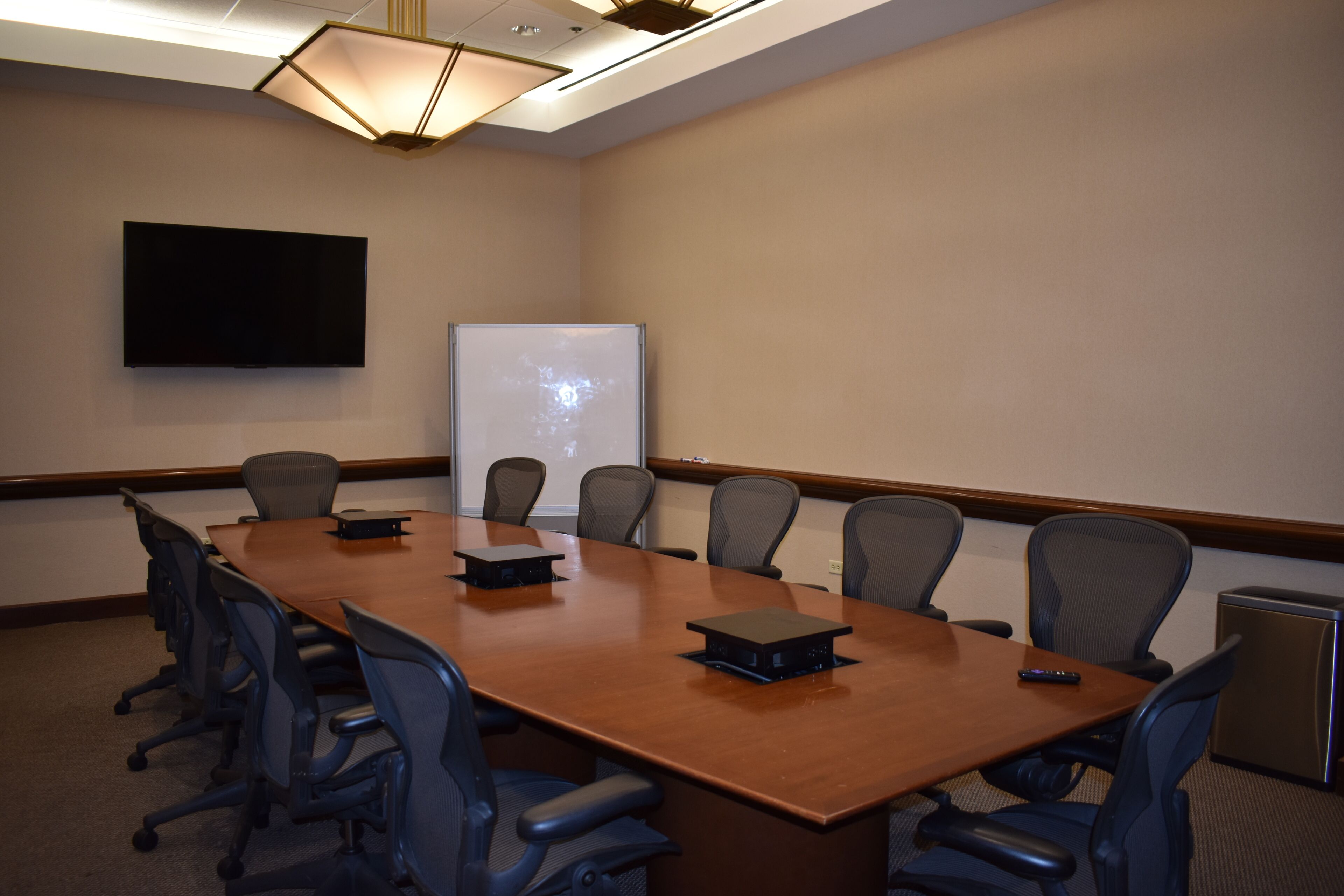 A conference room with a long table, chairs, a projector screen, and a flat-screen TV on the wall.