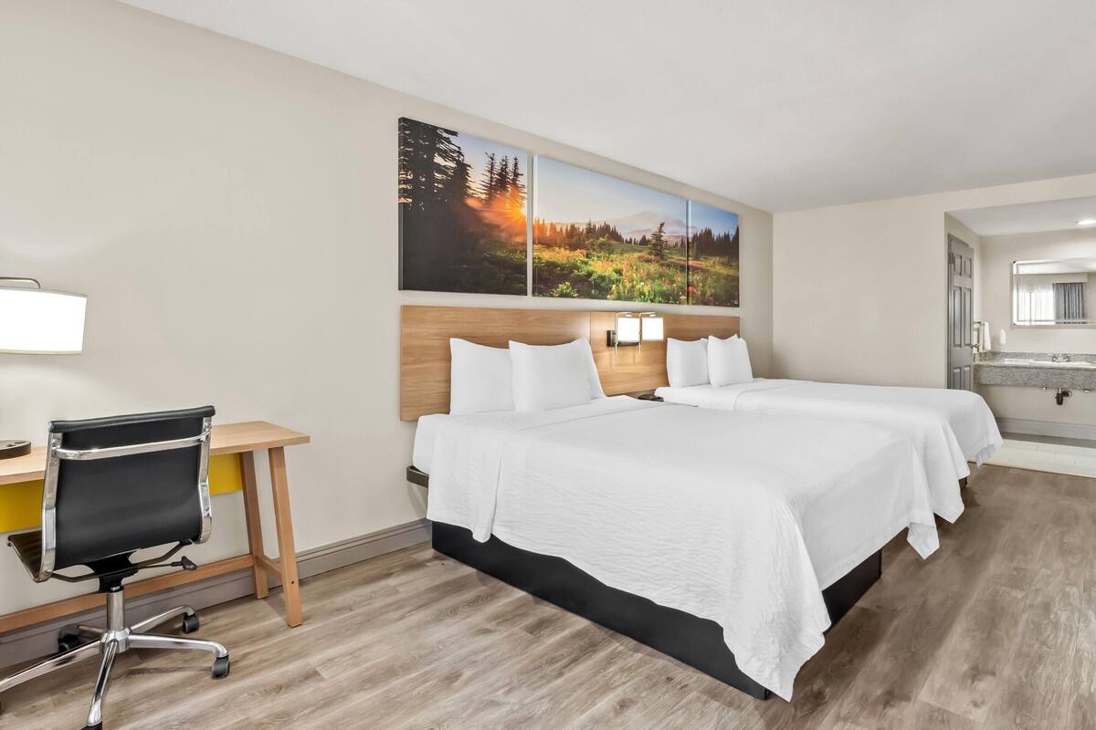 A hotel room with a wooden desk, two beds with white linens, a black office chair, and a large landscape painting on the wall.