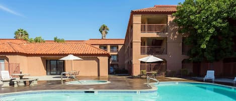 Una piscina con sillones de playa y sombrillas, rodeada por un edificio con balcones y palmeras.