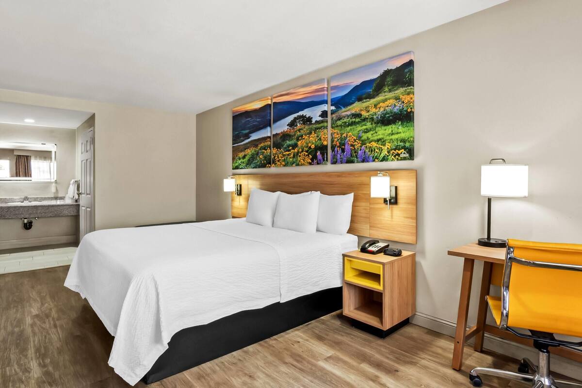 A hotel room with a large bed, a desk, a chair, a nightstand with a phone, and a painting above the bed.