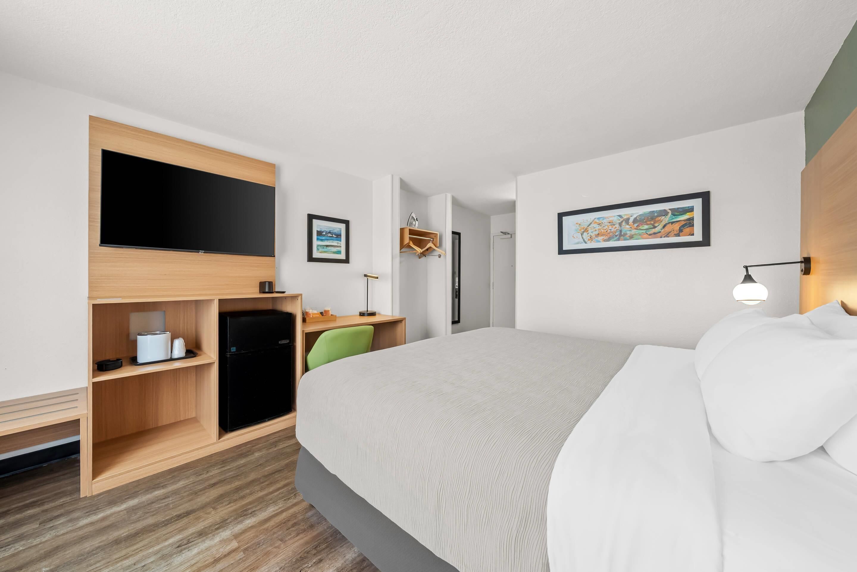 Quarto de hotel com cama, televisão, escrivaninha, cadeira e guarda-roupa.
