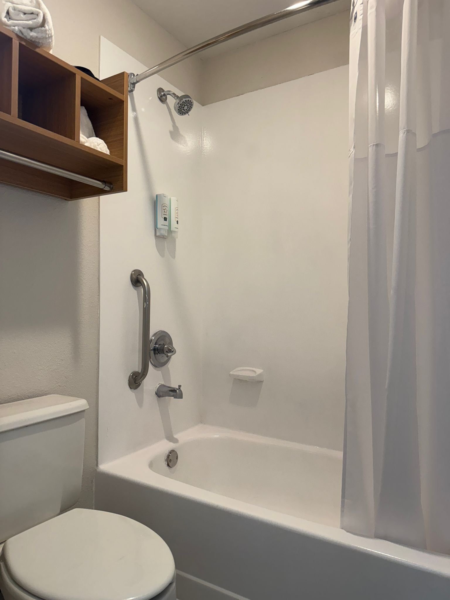 A bathroom with a white bathtub, a toilet, a shower curtain, and a wooden shelf above the toilet.