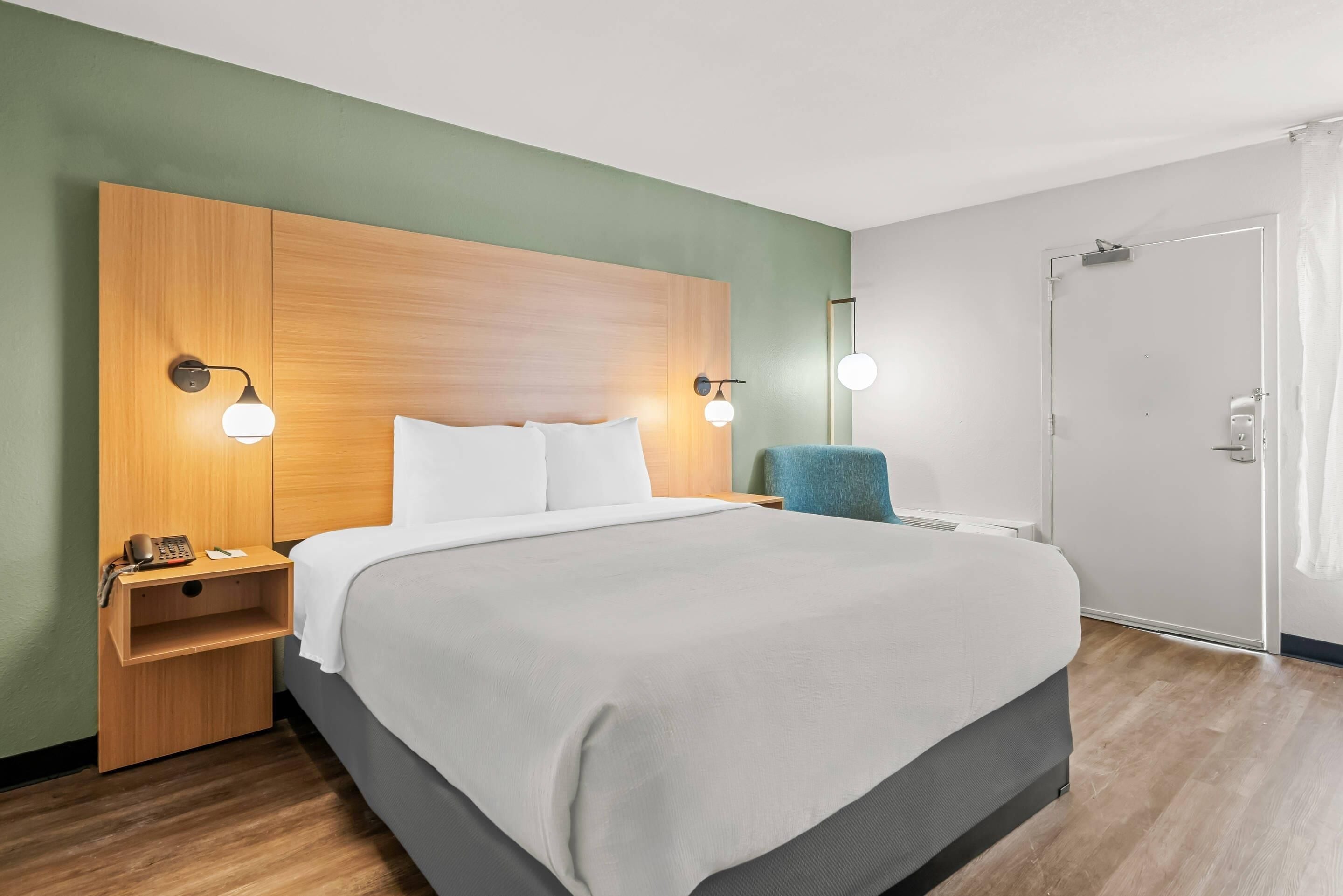 A hotel room with a large bed, wooden headboard, two bedside tables with lamps, a chair, and a door.