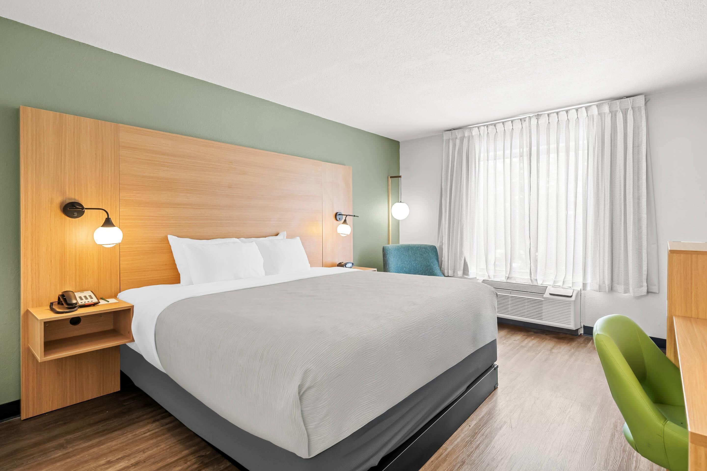 A hotel room with a large bed, bedside lamps, a green chair, a desk, and a window with curtains.