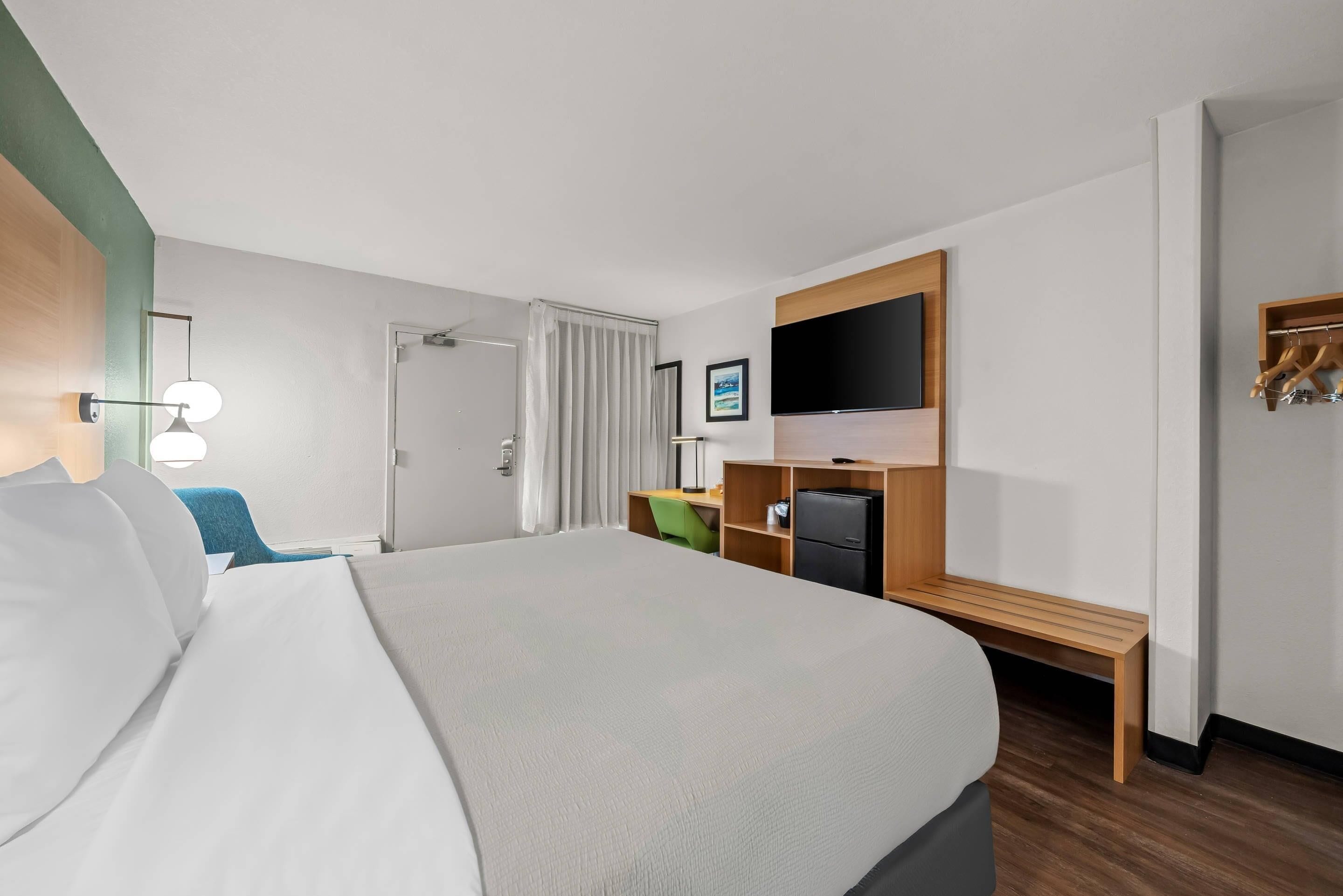 A hotel room with a large bed, a desk with a computer, a TV mounted on the wall, and a bench.