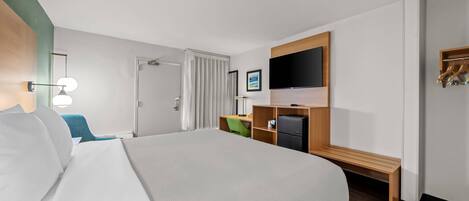 A hotel room with a large bed, a desk with a computer, a TV mounted on the wall, and a bench.