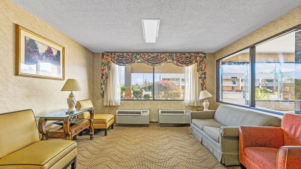 Ein Hotelzimmer mit beigeem Teppich, einem Sofa, einem roten Sessel, einem Tisch mit Lampe und einem fenster mit Vorhängen.