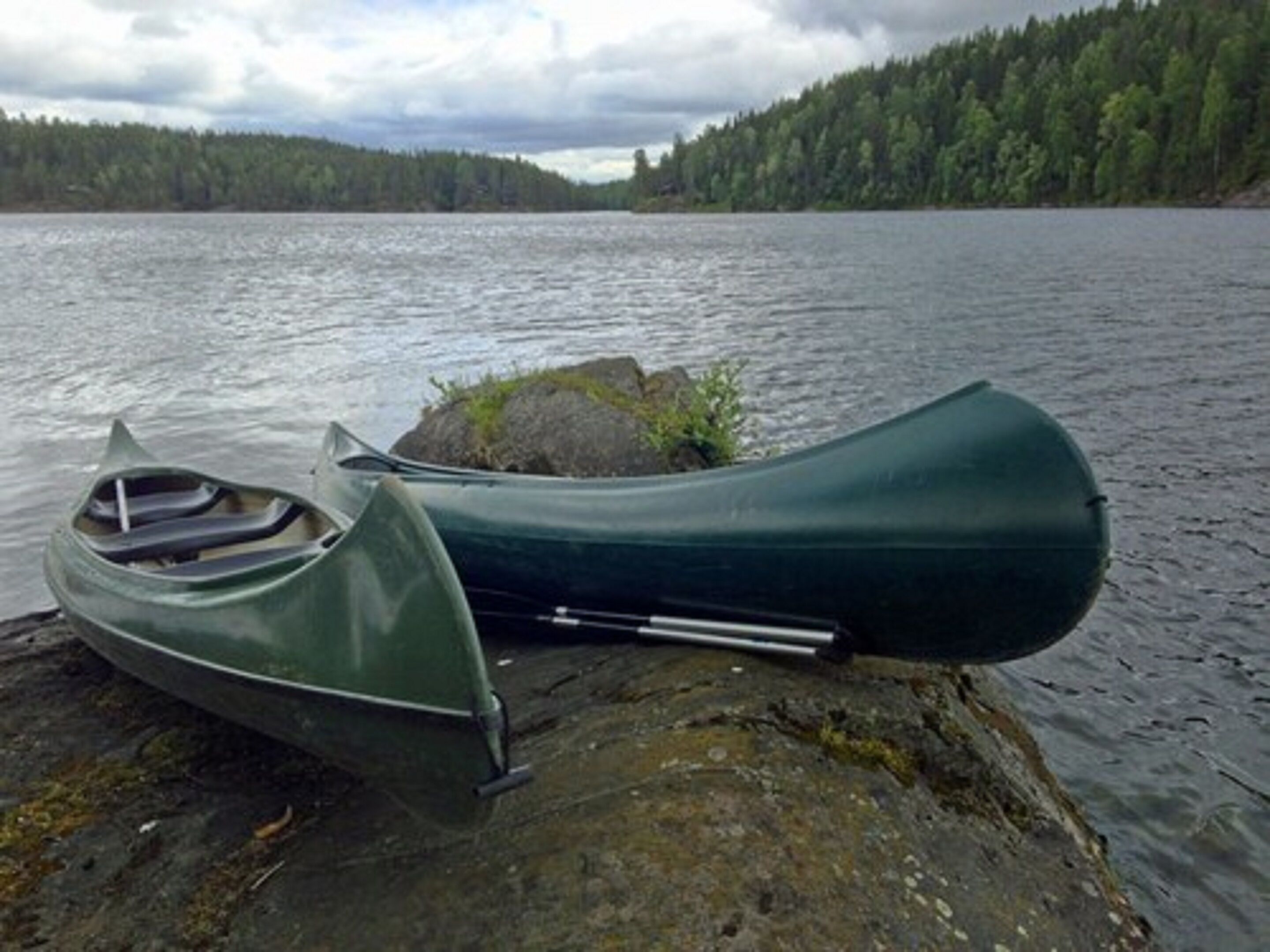Una canoa verde sobre una orilla rocosa junto a un lago, con un bosque al fondo.