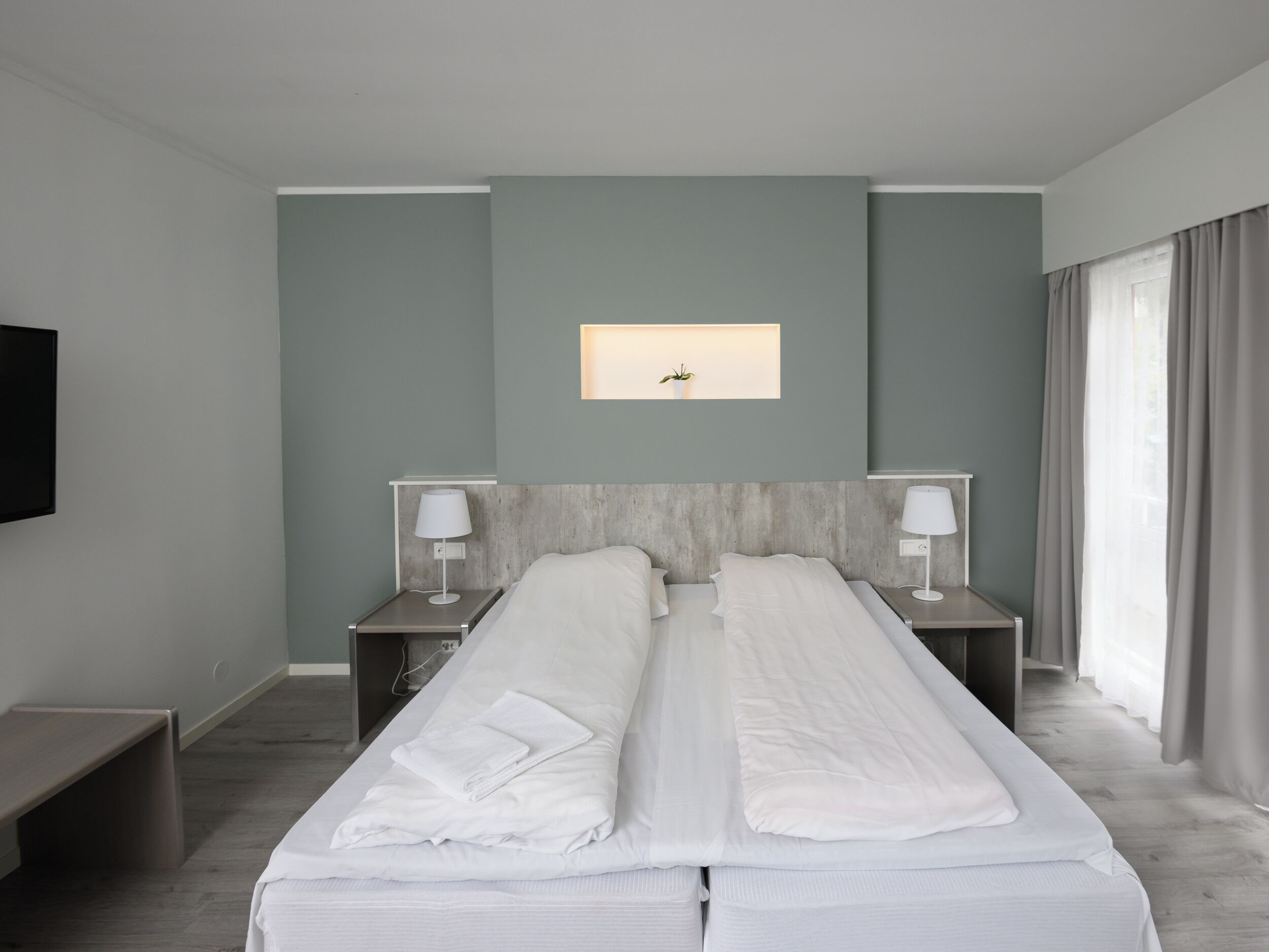 Ein modernes Hotelzimmer mit zwei Betten, einem Flachbildfernseher, einem Nachttisch mit Lampe und einem kleinen Fenster mit Vorhängen.