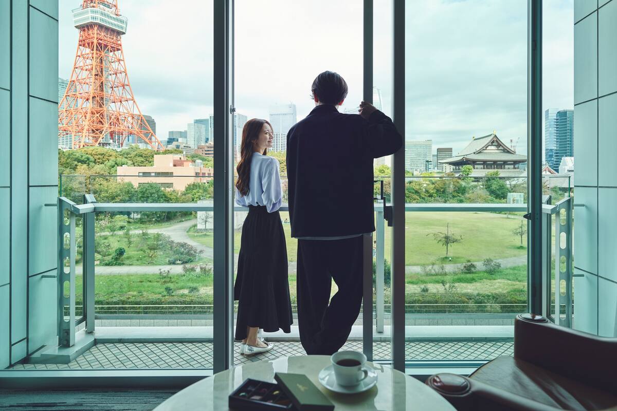 Un couple se tient près d’une grande fenêtre donnant sur un paysage urbain, avec une tour imposante et des bâtiments à l’architecture traditionnelle.