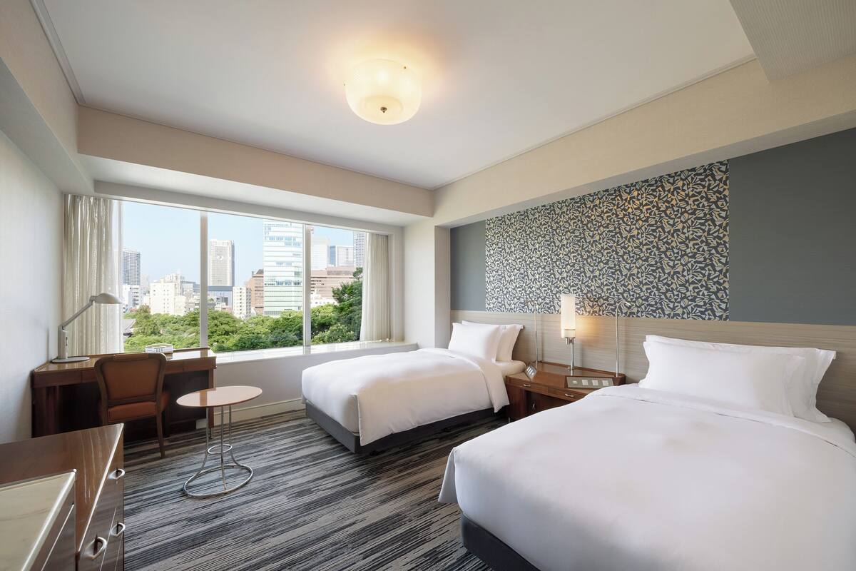 Une chambre d’hôtel avec deux lits, un bureau, une chaise, une petite table et une grande fenêtre donnant sur la ville.