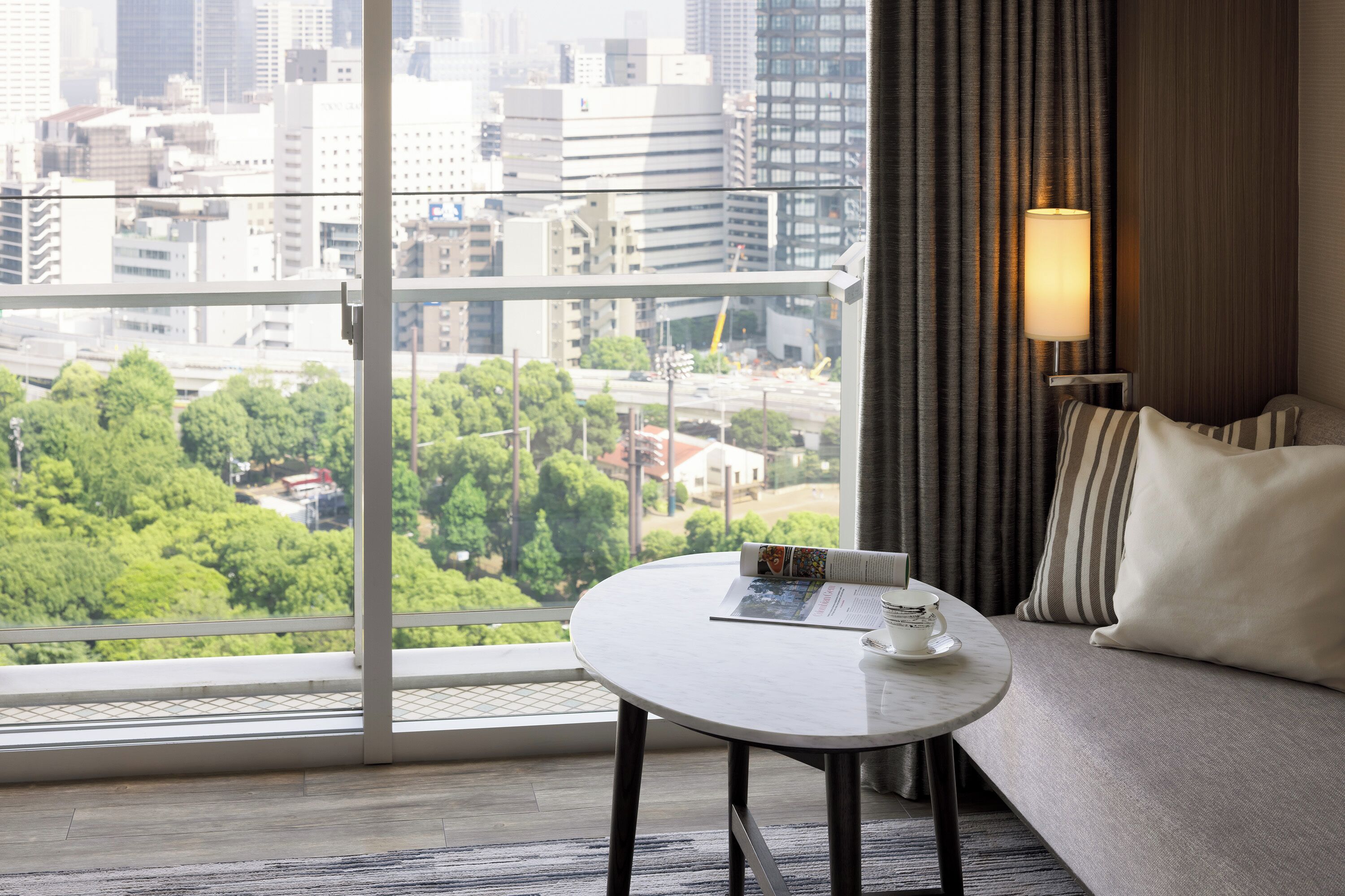 A hotel room with a large window offering a city view, a round table with a magazine and a cup, and a sofa with striped pillows.