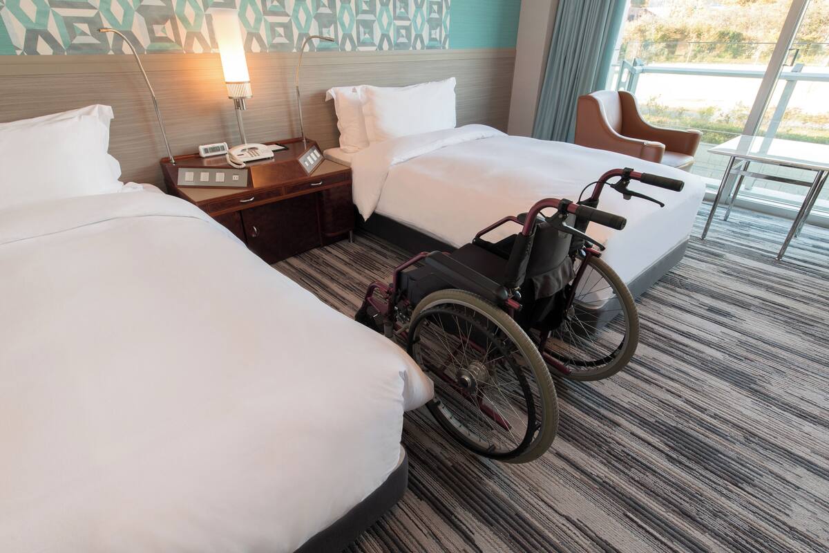 Une chambre d’hôtel avec deux lits, un fauteuil roulant, une table de chevet avec un téléphone et une fenêtre donnant sur un beau paysage.
