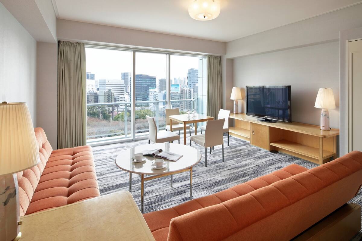 Une chambre d’hôtel moderne dotée d’une grande fenêtre, d’un canapé, d’une table à manger, d’un téléviseur et d’une lampe.