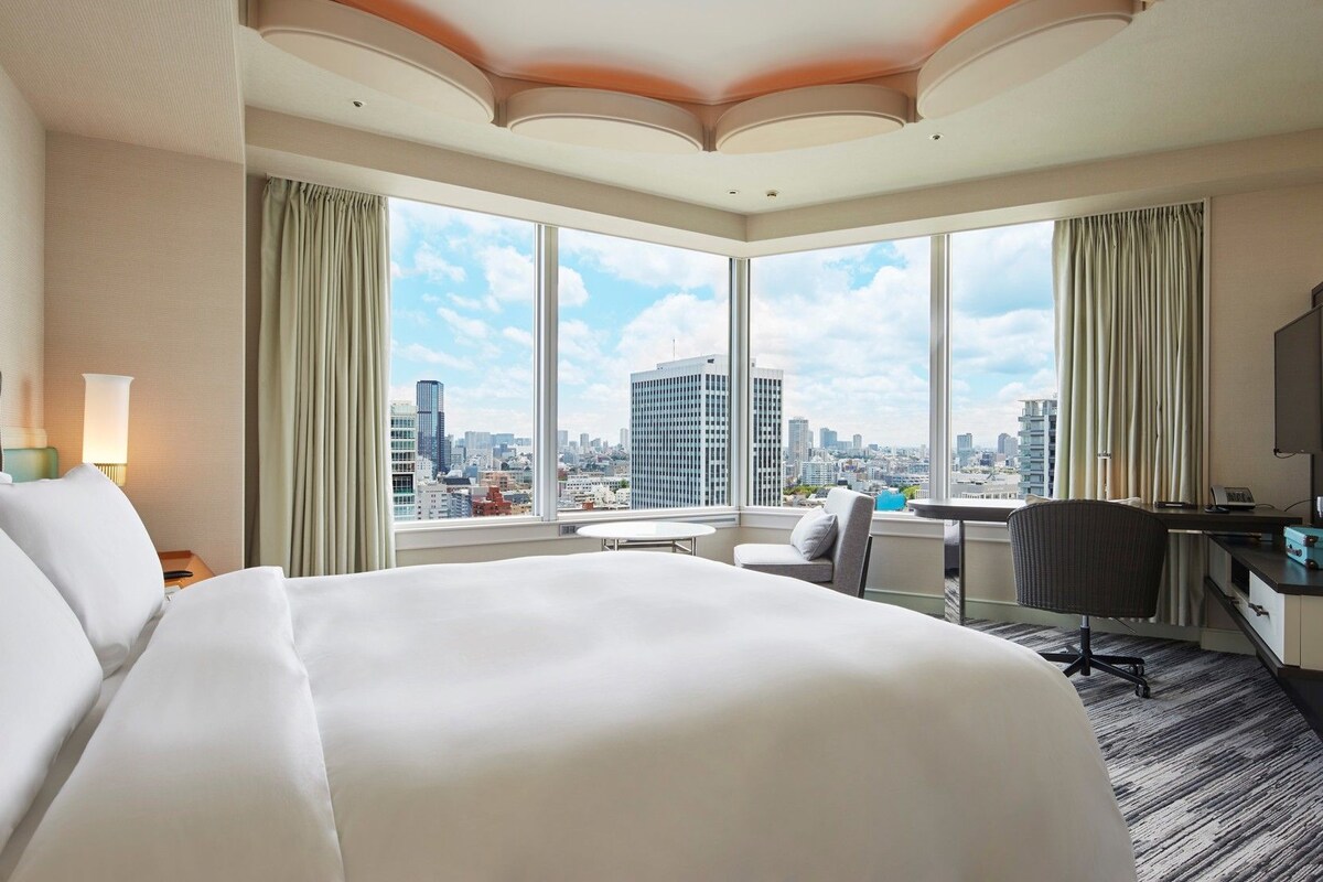 Une chambre d’hôtel avec un grand lit, un bureau, une chaise et une vue sur le paysage urbain grâce à de grandes fenêtres.