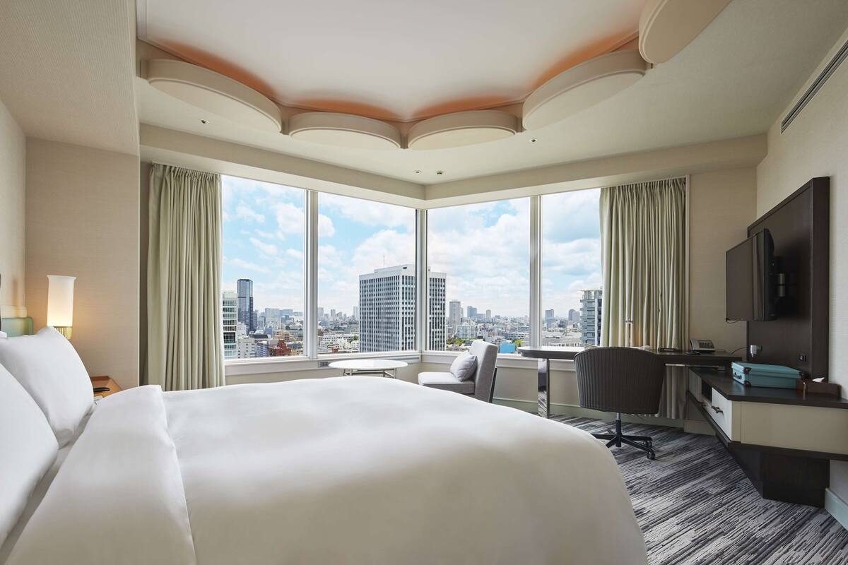 Une chambre d’hôtel avec un grand lit, un bureau, une chaise et une télévision. La chambre dispose de grandes fenêtres donnant sur la ville.
