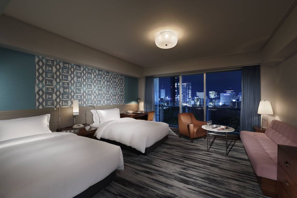 Une chambre d’hôtel avec deux lits, un canapé, une petite table et une vue sur le paysage urbain illuminé la nuit.