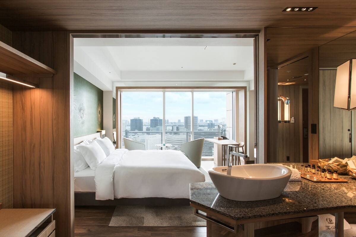 Une chambre d’hôtel moderne dotée d’un grand lit, d’une coiffeuse avec un grand lavabo et offrant une vue sur la ville grâce à de grandes fenêtres.