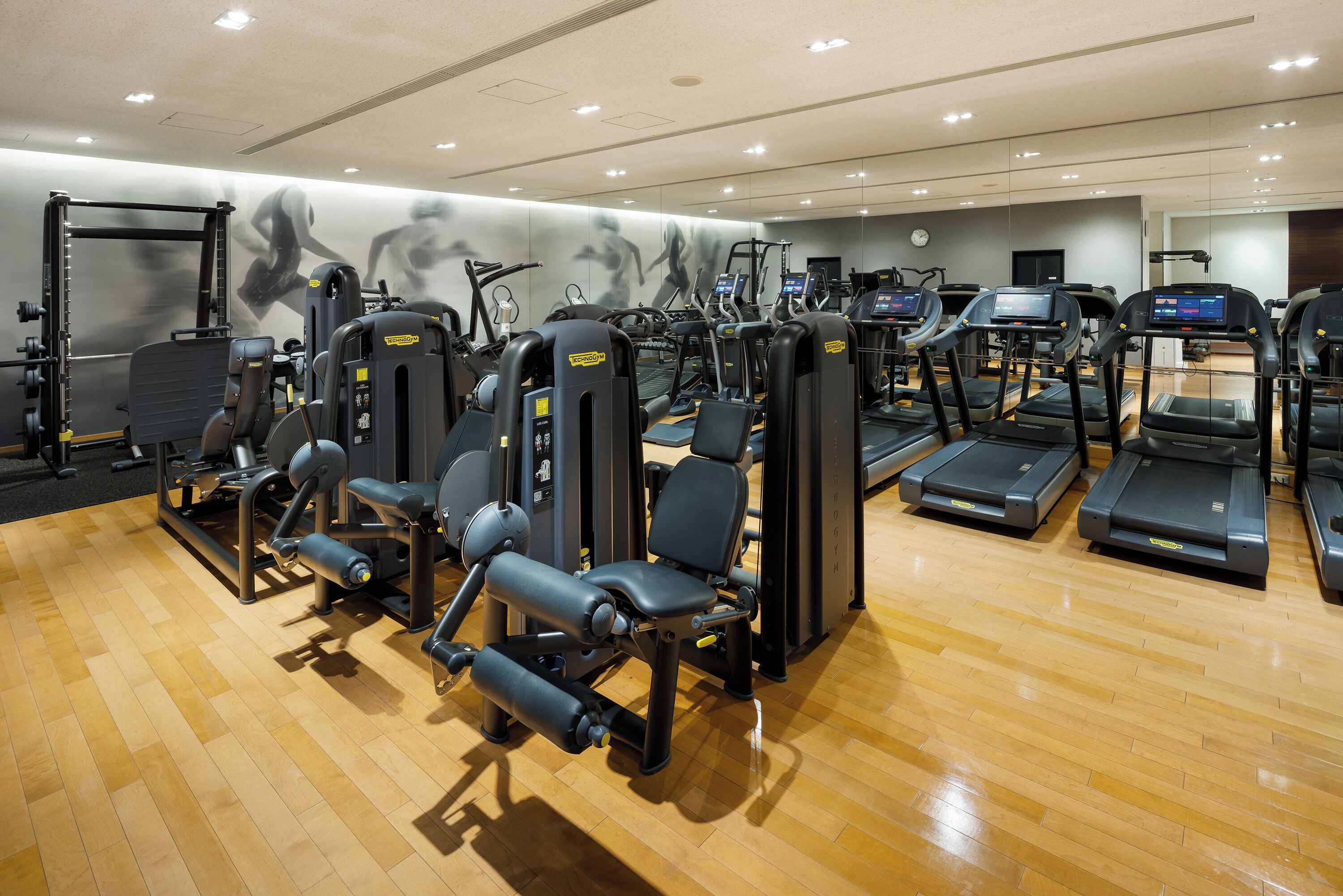 A gym with various exercise equipment, including treadmills and weight machines, on a wooden floor.