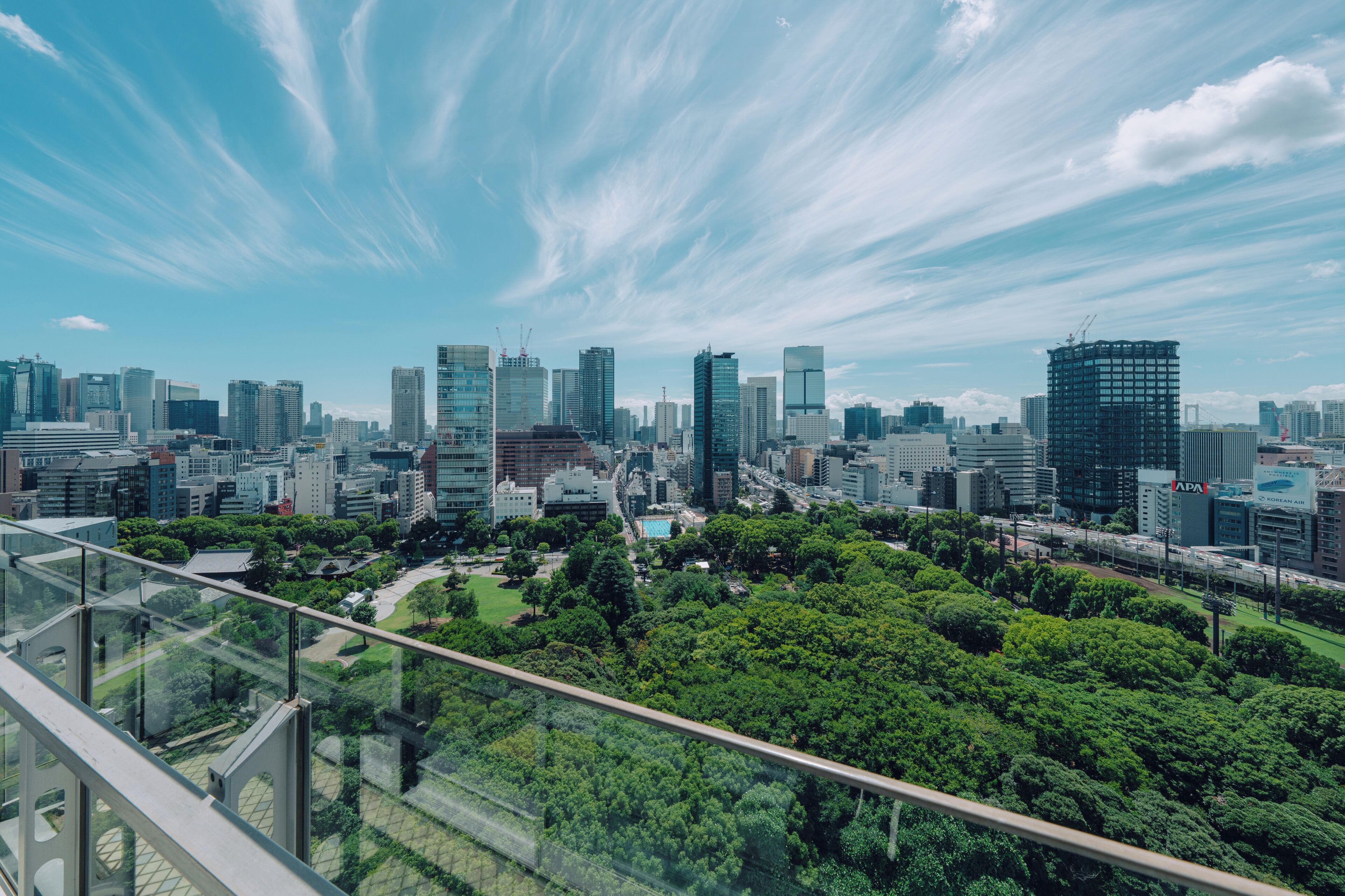 A cityscape with modern high-rise buildings, a park with dense green trees, and a clear blue sky with wispy clouds.