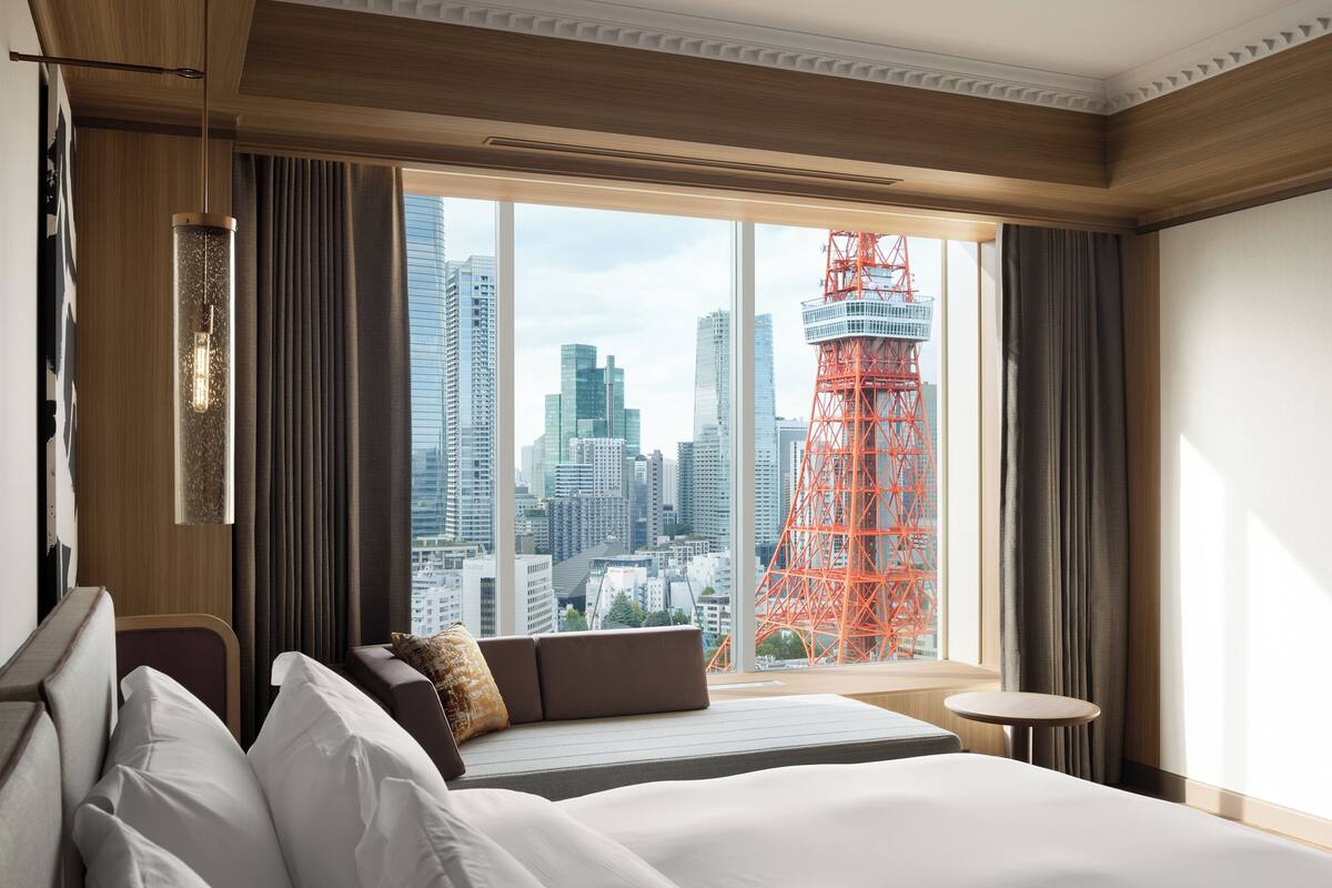 Une chambre d’hôtel dotée d’une grande fenêtre, d’un lit, d’un coin salon et offrant une vue sur le paysage urbain, notamment une tour rouge.