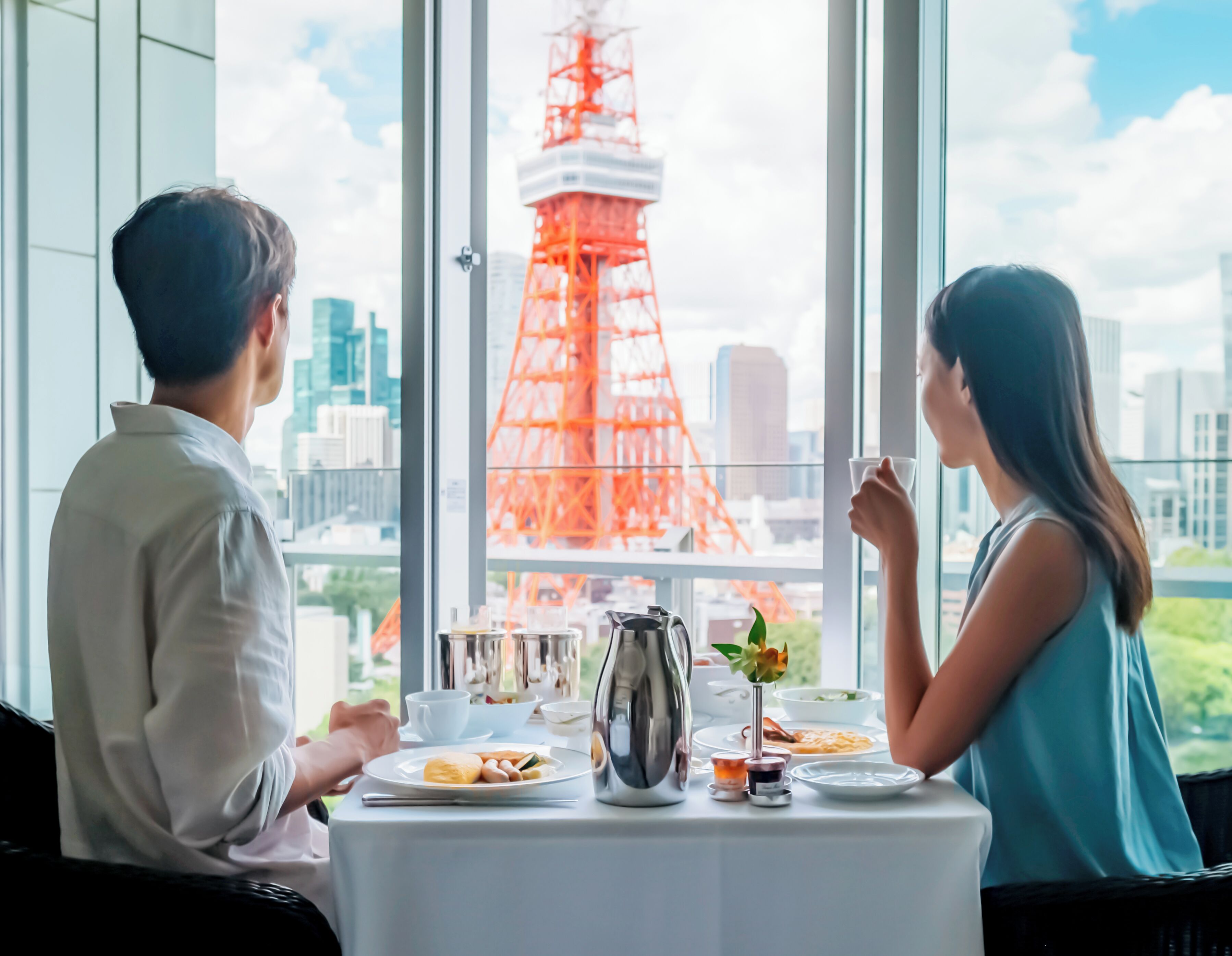 Uma mesa de jantar com pratos de comida, uma chaleira e uma xícara, em frente a uma janela com vista para uma torre vermelha e paisagem urbana.