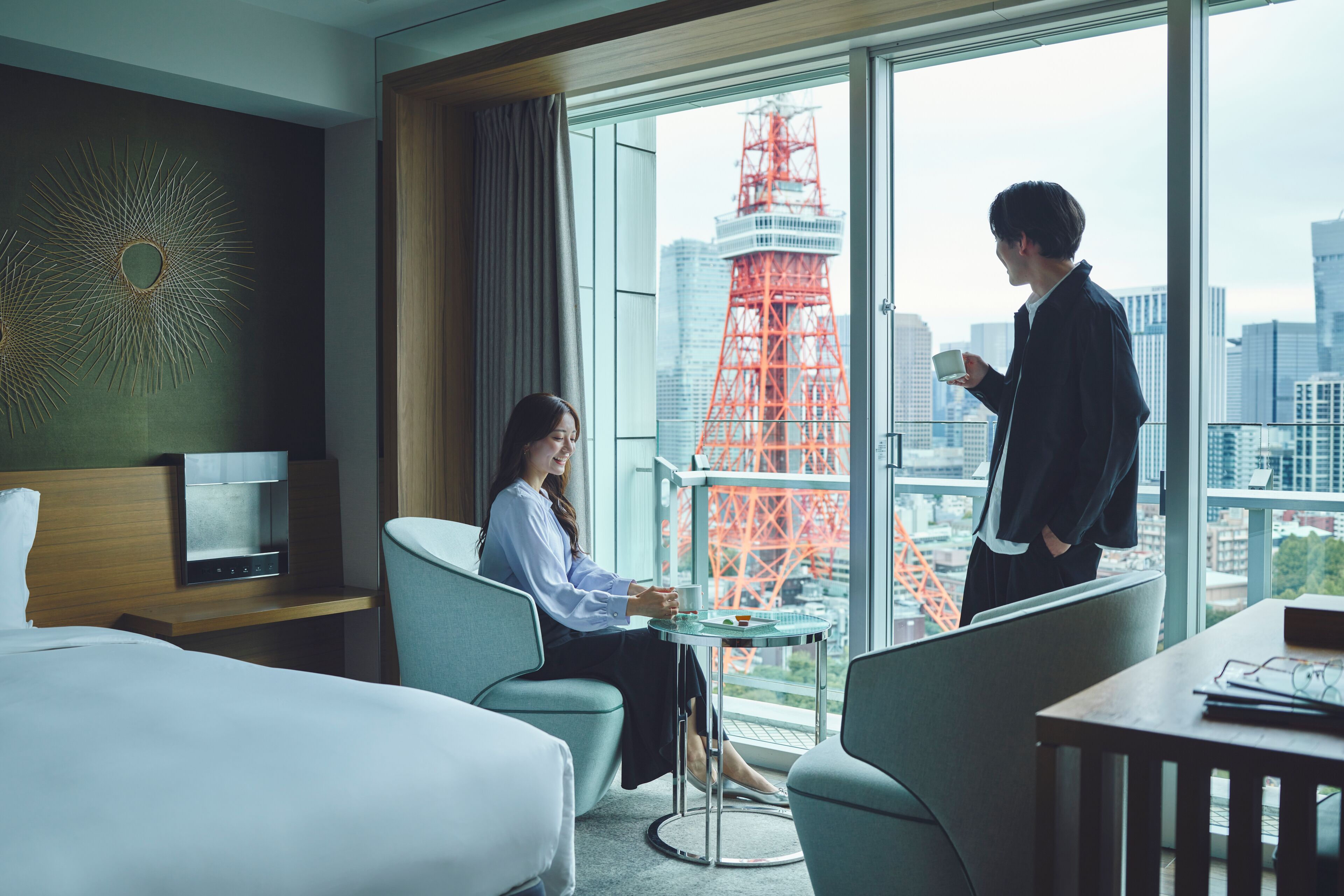 A hotel room with a bed, a desk, a chair, and a view of a cityscape including a red tower.