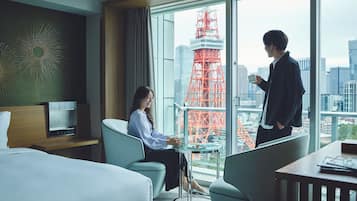 A hotel room with a bed, a desk, a chair, and a view of a cityscape including a red tower.