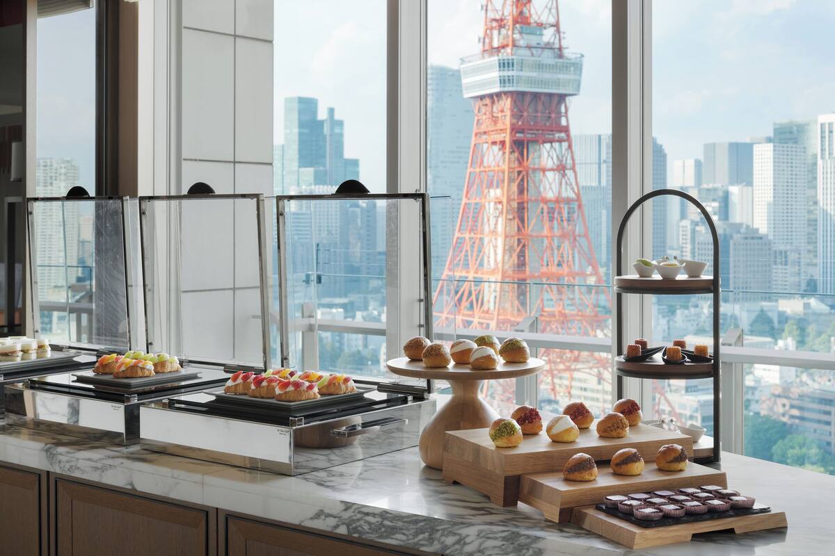 Un buffet proposant une variété de viennoiseries et de desserts, une grande fenêtre offrant une vue sur le paysage urbain et un immeuble, ainsi qu’un comptoir en marbre.