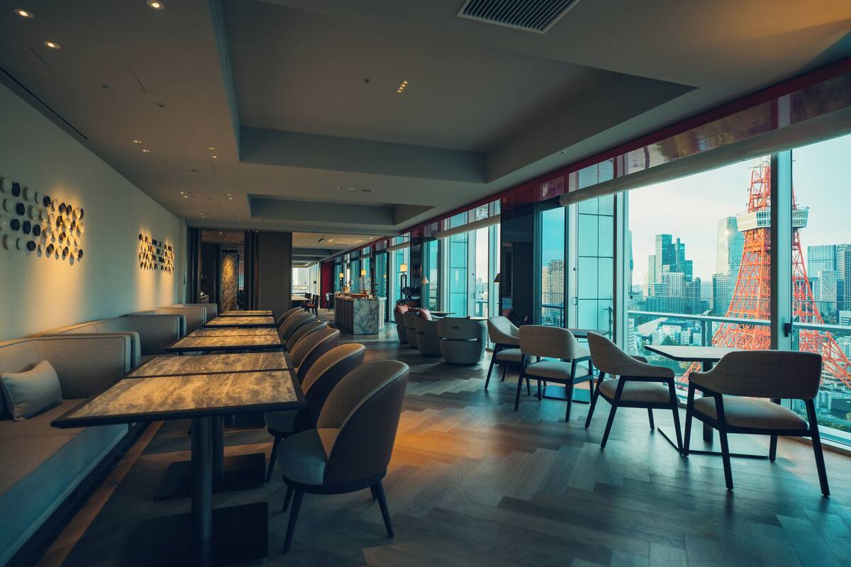 Un restaurant moderne avec de longues tables communes et des banquettes, de grandes fenêtres donnant sur la ville et une tour rouge distinctive en arrière-plan.
