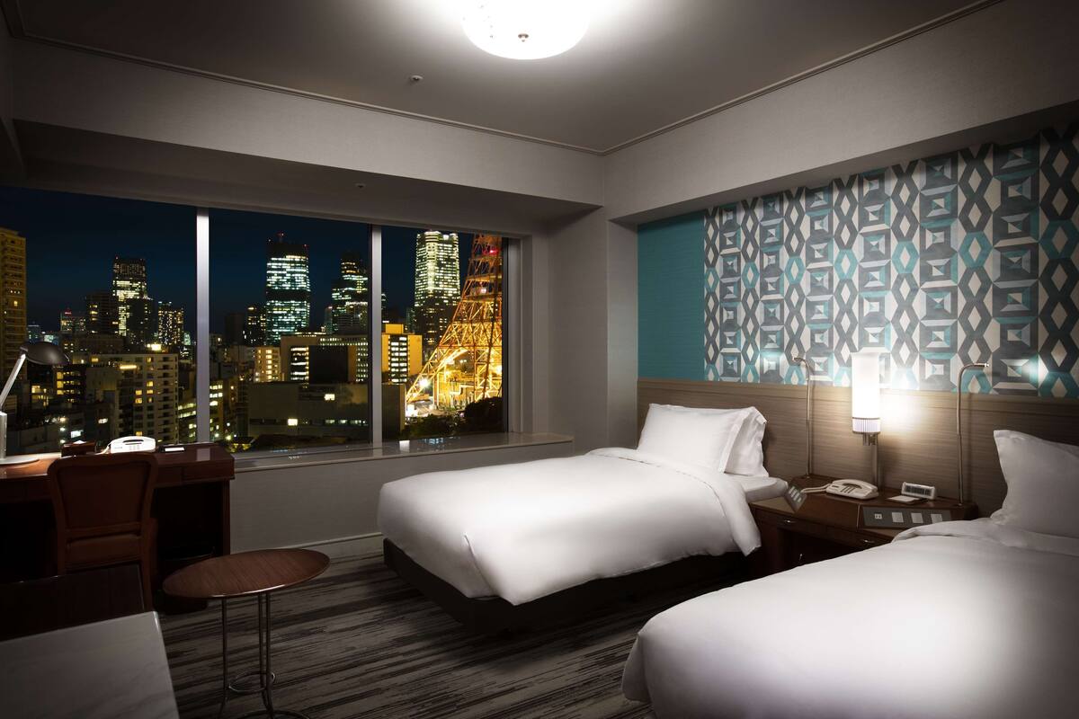 Une chambre d’hôtel avec deux lits, un bureau et une chaise. La chambre dispose d’une grande fenêtre donnant sur la ville.