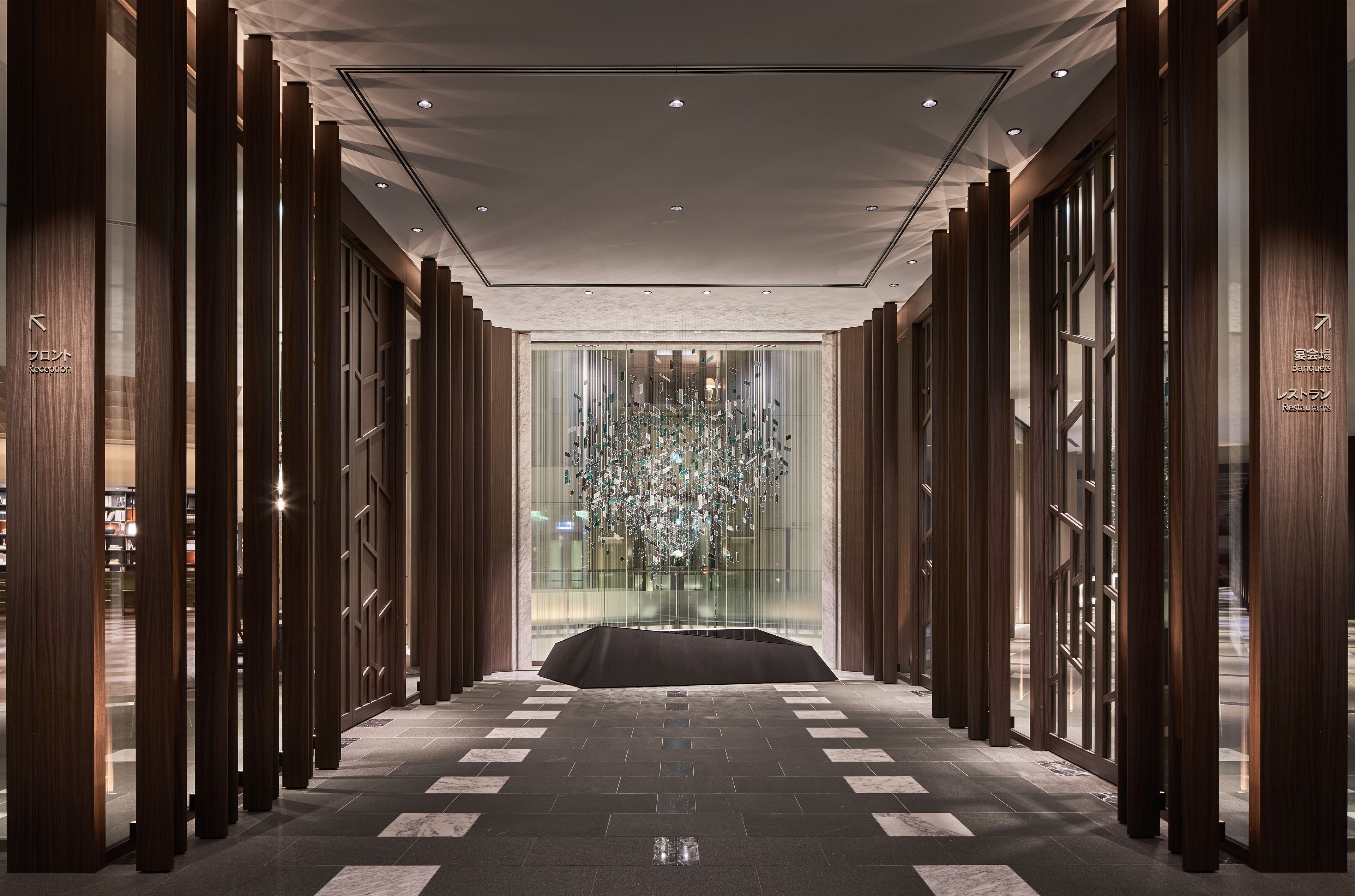 A modern hotel lobby with a central glass wall, wooden paneling, and a patterned floor.